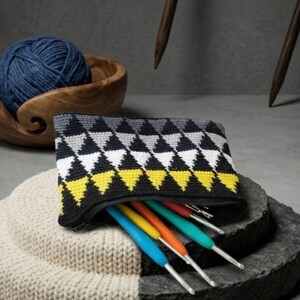 Peut inclure: Une trousse noire à fermeture éclair pour crochets, ornée d'un motif géométrique de triangles jaunes, blancs, gris et noirs. La trousse est ouverte, révélant plusieurs crochets aux manches colorées. Une pelote de laine bleue est dans un bol en bois.