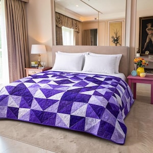 May include: A geometric quilt in shades of purple and white covers a bed with white pillows. The quilt features a repeating pattern of triangles and squares. The bed is in a well-lit room with a neutral color scheme.