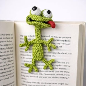 May include: A whimsical, handmade crochet frog bookmark. The green frog has large white eyes with black pupils and a red tongue sticking out. It is placed in an open book, adding a playful touch to reading.