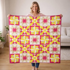 May include: A colorful quilt with a star pattern in shades of pink, yellow, and beige. The quilt features a repeating geometric design with a pink border. The quilt is held up, showcasing its size and design.