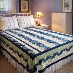 May include: A blue and white patterned quilt on a bed with white ruffled pillows. The quilt features a geometric design with a green border and a white bed skirt. A lamp with a stained glass shade sits on a side table.