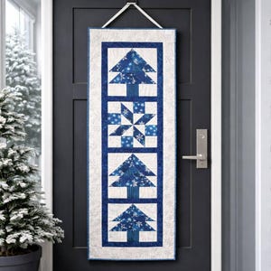 May include: A rectangular, quilted wall hanging with a winter theme. The design features blue Christmas trees and a snowflake pattern on a white background, framed by a blue border. It is suspended by a white ribbon.