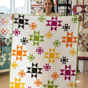 May include: A handmade quilt with a white background and colorful star patterns in black, orange, green, yellow, and purple. The quilt has a green border and is displayed in a well-lit room, showcasing the craftsmanship of the textile art.