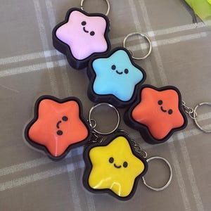 May include: A collection of star-shaped keychains in various colors: pink, blue, orange, and yellow. Each keychain has a black border and a smiling face design. The keychains are attached to silver key rings.