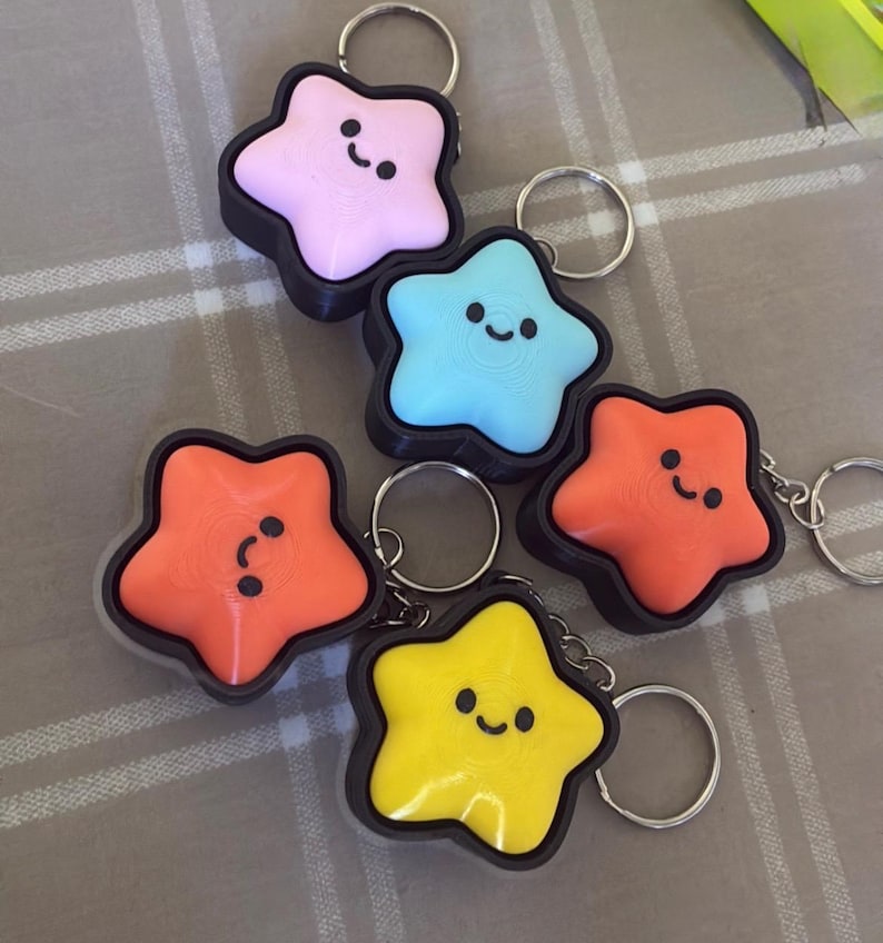 May include: A collection of star-shaped keychains in various colors: pink, blue, orange, and yellow. Each keychain has a black border and a smiling face design. The keychains are attached to silver key rings.
