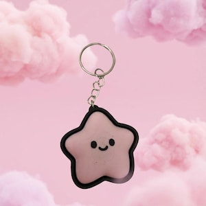 May include: A pink star-shaped keychain with a black outline and a smiling face. The keychain is attached to a silver ring and chain. The background features pink clouds against a pink backdrop.