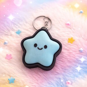 May include: A light blue star-shaped keychain with a smiling face and a dark brown border. The keychain is attached to a silver metal ring. The background is a soft, colorful blend of pink, blue, and yellow.