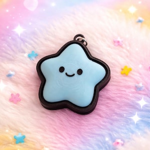 May include: A light blue star-shaped charm with a black border and a smiling face. The charm is on a fluffy, multi-colored surface with star and heart-shaped embellishments. The charm has a small loop for attaching to a keychain or necklace.