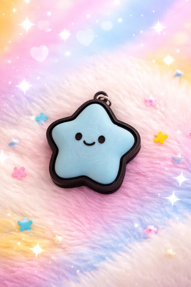 May include: A light blue star-shaped charm with a black border and a smiling face. The charm is on a fluffy, multi-colored surface with star and heart-shaped embellishments. The charm has a small loop for attaching to a keychain or necklace.