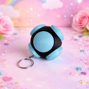 Könnte beinhalten: Ein schwarzer und hellblauer kugelförmiger Schlüsselanhänger. Der Schlüsselanhänger hat einen silbernen Ring und befindet sich vor einem rosa Hintergrund mit einem Regenbogen, Blumen und sternförmigem Konfetti.