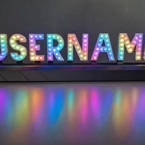 May include: A decorative sign spelling "USERNAME" in white block letters, each lit with multi-coloured LED lights. The sign rests on a black base, creating a rainbow reflection on the surface beneath it.
