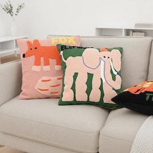 Könnte beinhalten: Eine Sammlung dekorativer Kissen mit Tiermotiven. Ein Kissen zeigt einen orangefarbenen Fuchs auf rosa Hintergrund, ein anderes einen rosa Elefanten auf grünem Grund und ein drittes einen Tiger. Die Kissen liegen auf einem neutralfarbenen Sofa.