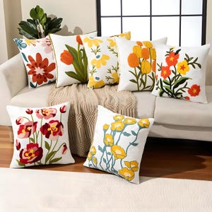 Könnte beinhalten: Eine Auswahl weißer Zierkissen mit farbenfrohen Blumenstickereien. Die Kissen zeigen verschiedene Blumendesigns in Orange, Gelb, Rot und Rosa, mit grünen Blättern. Die Kissen sind auf einem cremefarbenen Sofa angeordnet.