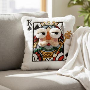May include: White plush decorative pillow featuring an embroidered King of Spades playing card design. The king has a fluffy mustache and eyebrows, a crown, and holds a scepter. The pillow is on a gray couch with a white blanket.