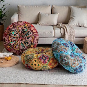 Könnte beinhalten: Drei runde Bodenkissen mit farbenfrohen, gemusterten Stoffen. Die Kissen sind in Rot-, Blau- und Gelbtönen gehalten und weisen aufwendige Designs auf. Sie liegen auf einem weißen Teppich vor einem beigen Sofa.