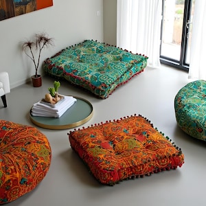 May include: Several floor cushions with colorful, patterned fabric are displayed. The cushions are square and round, with a buttoned center and a pom-pom trim. The dominant colors are teal, orange, and red, with intricate floral and paisley designs.