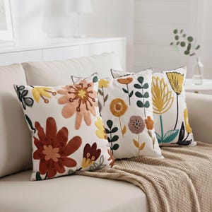 May include: Three embroidered floral cushions in yellow, orange, and brown on a white background. The cushions are placed on a beige sofa with a matching throw. The floral patterns feature various flower types and leaf details, adding a touch of nature.