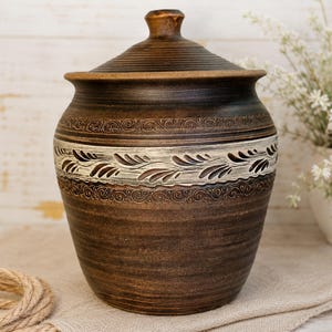 May include: A brown ceramic jar with a lid, featuring a decorative band of white and brown leaf patterns. The jar has a textured surface and a small, rounded knob on top. The jar is sitting on a beige cloth.