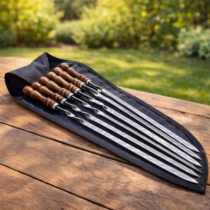Op de afbeelding: Een set roestvrijstalen spiesen met houten handvatten, netjes gerangschikt in een zwarte draagtas. De spiesen zijn lang en slank, perfect om te grillen. De houten handvatten zijn diep bruin van kleur en bieden een comfortabele grip.