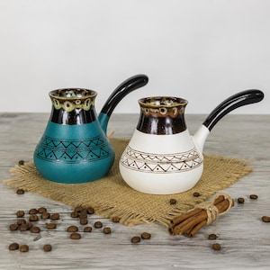 May include: Two ceramic coffee pots with long black handles. One pot is teal with a dark brown neck and a geometric pattern, while the other is white with a dark brown neck and a matching geometric pattern. Both feature a speckled rim design.