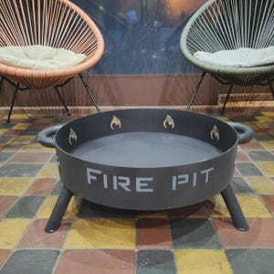 Peut inclure: Un foyer extérieur gris foncé avec l'inscription "FIRE PIT" en lettres blanches. Le foyer a un design circulaire avec trois pieds et deux poignées. Le bord comporte des découpes de flammes.
