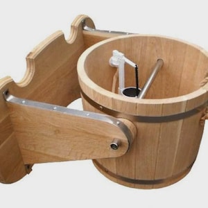 Puede incluir: Un cubo de sauna de madera con una banda de metal y un asa de cuerda. El cubo está montado en un marco de madera con un borde decorativo. Un mecanismo blanco y negro está dentro del cubo.