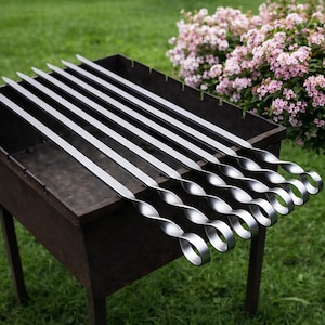 Könnte beinhalten: Ein Satz Edelstahlspieße mit gedrehten Griffen, die auf einem dunklen Metallgrill liegen. Die Spieße sind parallel angeordnet, mit einem grünen Rasen und rosa blühenden Büschen im Hintergrund. Ideal zum Grillen.