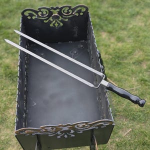 Könnte beinhalten: Ein schwarzer Metallgrill mit dekorativen Ausschnitten und einer Edelstahl-Grillgabel mit schwarzem Griff. Der Grill ist rechteckig und steht auf einer grasbewachsenen Oberfläche.