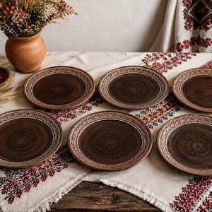 May include: A set of six round brown ceramic plates featuring a textured, embossed pattern along the outer rim. Each plate has a dark, mottled finish with a smooth center, showcasing a consistent handcrafted appearance across the entire collection.