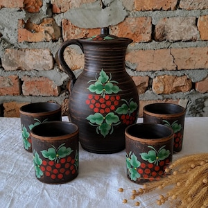 Pode incluir: Um jarro de cerâmica castanho-escuro com tampa e quatro chávenas a condizer, todas decoradas com cachos de bagas vermelhas e folhas verdes pintadas à mão. O jarro apresenta uma pega curva e estrias horizontais à volta do pescoço.