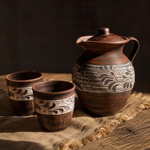 Può includere: Una brocca rustica in ceramica marrone con coperchio e manico, insieme a due tazze abbinate. La ceramica presenta un motivo floreale decorativo bianco e marrone al centro. Il set è esposto su un panno di juta.