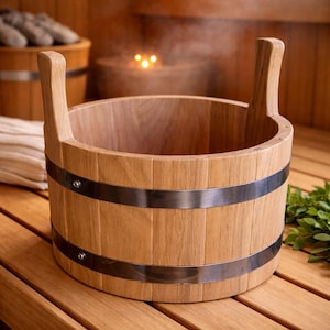 Puede incluir: Un cubo de sauna de madera marrón claro con dos asas verticales. El cubo se sujeta con dos bandas de metal plateado. El cubo de sauna está sobre una superficie de madera.