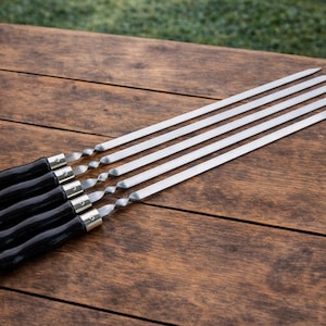 Puede incluir: Un juego de seis brochetas de acero inoxidable con mangos de madera negros. Las brochetas están dispuestas en diagonal sobre una superficie de madera. Las brochetas están diseñadas para asar a la parrilla.