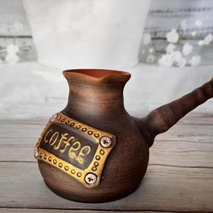 May include: A handmade brown ceramic coffee pot with a long spout and a decorative gold-edged plaque that reads "coffee". The pot has a rustic, artisanal appearance, perfect for brewing coffee.