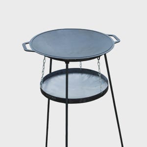 Puede incluir: Un soporte de cocina exterior de metal negro con una superficie de cocción redonda y un estante inferior. El soporte tiene tres patas y está diseñado para asar a la parrilla o cocinar al aire libre. La superficie de cocción tiene asas.