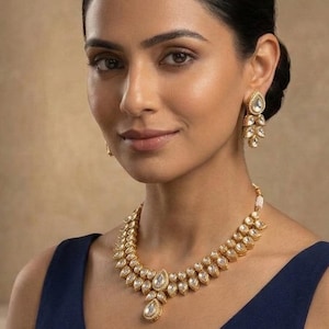 May include: A gold necklace and earrings set. The necklace features a double strand design with a large, pear-shaped pendant. The earrings have a similar design. The woman is wearing a navy blue dress with a V-neck.