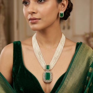May include: A multi-strand pearl necklace with a large emerald pendant and matching square emerald earrings. The pendant and earrings are framed with a silver-colored metal and small clear stones. The woman is wearing a dark green velvet top.