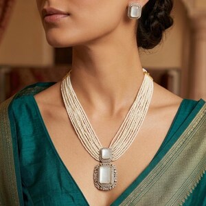 May include: A pearl necklace with a large, square pendant and matching earrings. The pendant and earrings have a white center stone surrounded by a decorative border. The necklace features multiple strands of small pearls.