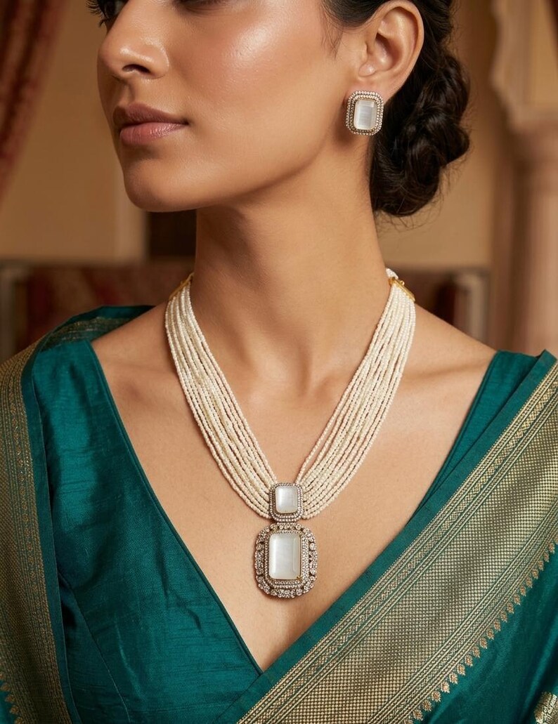 May include: A pearl necklace with a large, square pendant and matching earrings. The pendant and earrings have a white center stone surrounded by a decorative border. The necklace features multiple strands of small pearls.