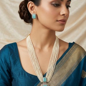 May include: A woman wearing a multi-strand pearl necklace with a large rectangular pendant and matching square earrings. The pendant and earrings feature a light blue gemstone surrounded by a decorative border. The woman is wearing a teal blue top.