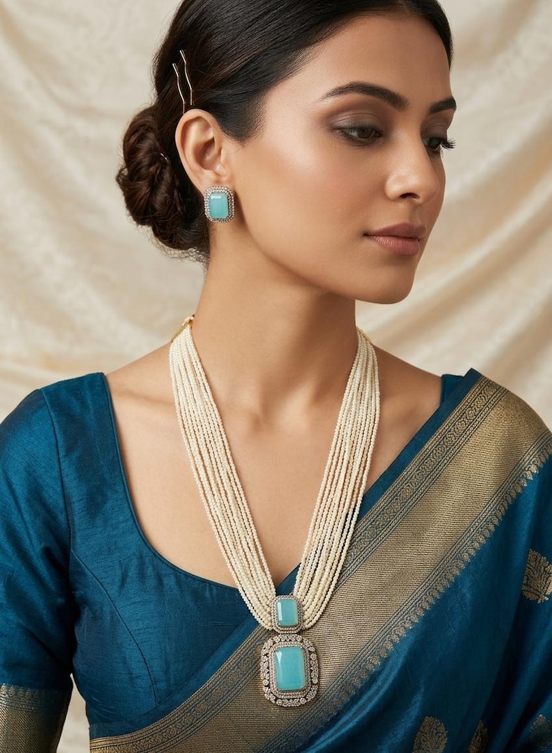 May include: A woman wearing a multi-strand pearl necklace with a large rectangular pendant and matching square earrings. The pendant and earrings feature a light blue gemstone surrounded by a decorative border. The woman is wearing a teal blue top.