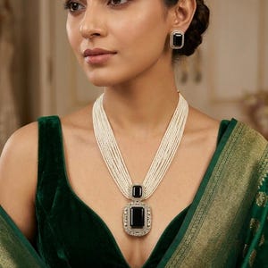 May include: A multi-strand pearl necklace with a large, rectangular black pendant and matching square earrings. The pendant and earrings are framed with a silver-colored setting. The woman is wearing a dark green velvet top and a green and gold patterned shawl.