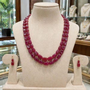 May include: A double-strand necklace featuring polished, oval-shaped deep red ruby beads, accompanied by a pair of matching drop earrings, each consisting of two stacked ruby beads suspended from a small gold-toned metal hook fixture.