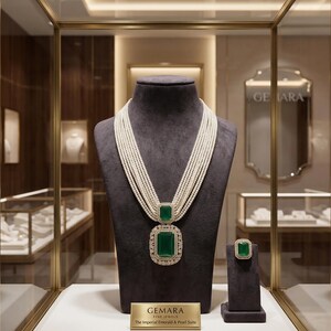 May include: A luxurious jewelry display featuring a pearl necklace with a large emerald pendant and matching ring. The necklace has multiple strands of pearls. The emeralds are set in a gold frame and surrounded by smaller diamonds. The display includes the text "GEMARA FINE JEWELS".