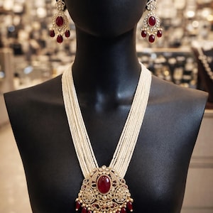 Puede incluir: Un collar de perlas de múltiples hilos con un colgante grande y ornamentado con un rubí ovalado central y pequeños acentos de rubí. Los pendientes a juego de rubí y oro completan el conjunto, exhibido en un maniquí negro.