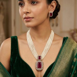 May include: A woman wearing a necklace and earrings set. The necklace features a large, rectangular, red gemstone pendant surrounded by small, clear stones, suspended from multiple strands of white beads. The earrings have matching red gemstones. The woman is wearing a dark green velvet top.