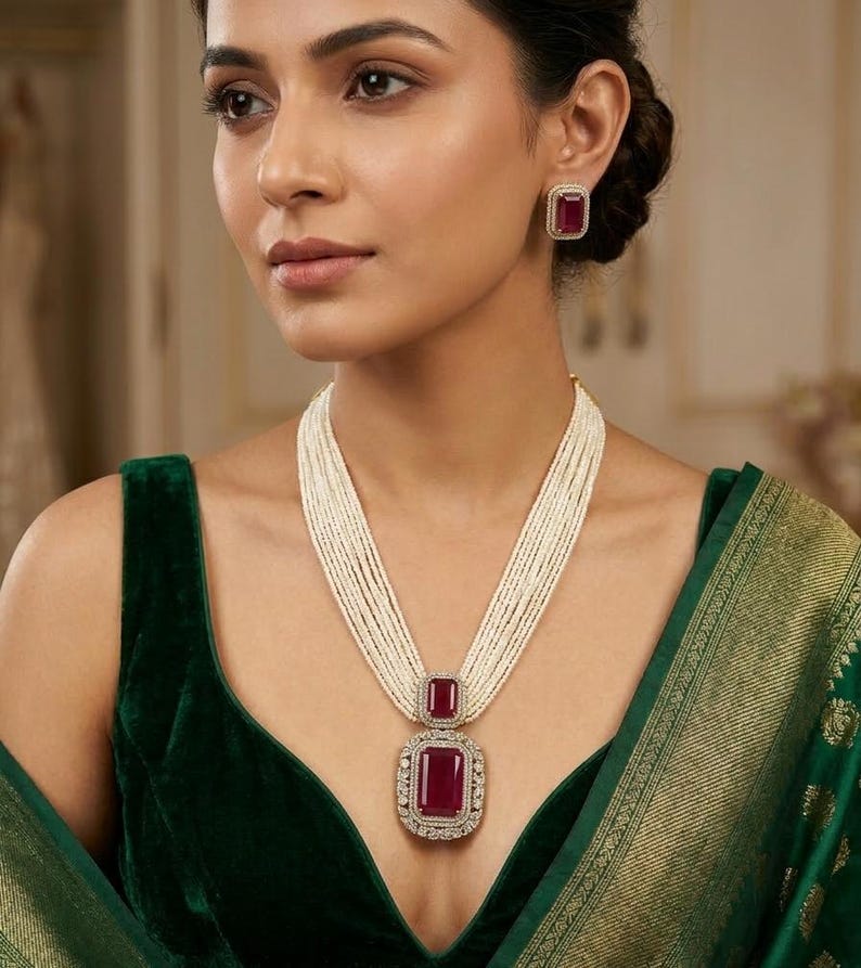 May include: A woman wearing a necklace and earrings set. The necklace features a large, rectangular, red gemstone pendant surrounded by small, clear stones, suspended from multiple strands of white beads. The earrings have matching red gemstones. The woman is wearing a dark green velvet top.