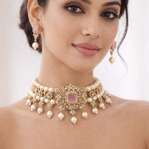 May include: A gold-toned necklace and matching earrings set. The necklace features a central pink square stone, surrounded by an ornate design and accented with pearls. The earrings have a similar design with a pink square stone and a dangling pearl.