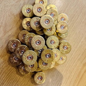 Può includere: Un gruppo di bossoli di cartucce per fucile calibro 12 in ottone. Ogni bossolo è marchiato con il marchio "ELEY" e il numero "12". I bossoli sono disposti su una superficie di legno.