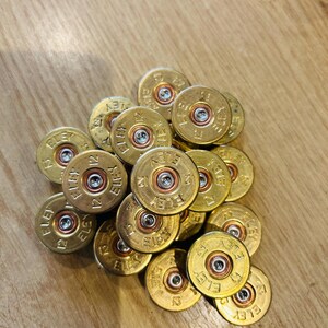 Può includere: Un mucchio di bossoli di cartucce per fucile in ottone dorato con la scritta "ELEY 12". I bossoli, con centri argentati, sono disposti su una superficie di legno. Ogni bossolo misura circa 2 cm di diametro.